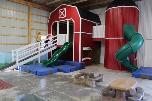 Indoor play area featuring a red barn structure with slides, climbing areas, and soft play mats.
