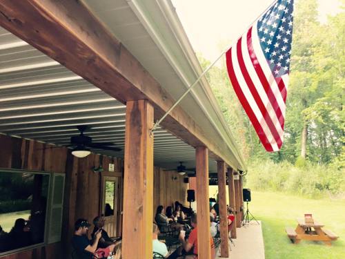 A group of people seated under a covered porch, with an American flag waving nearby and trees in the background.