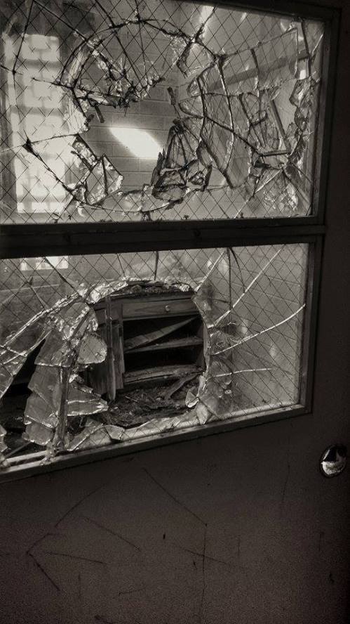 A close-up of a broken glass door with shattered panes and a hole, revealing a dark interior space.