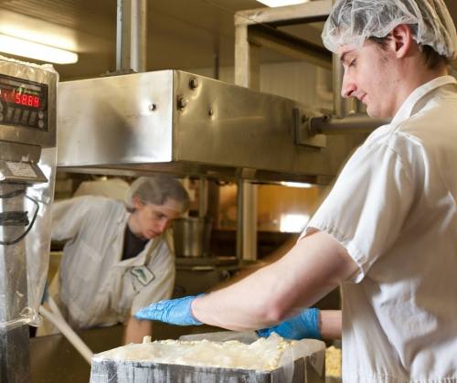 Two workers in a food processing facility, one measuring ingredients and the other mixing in the background.