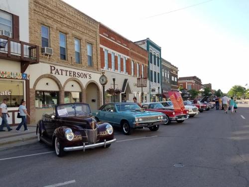 A row of classic cars parked along a historic downtown street with people walking by and shops in the background.