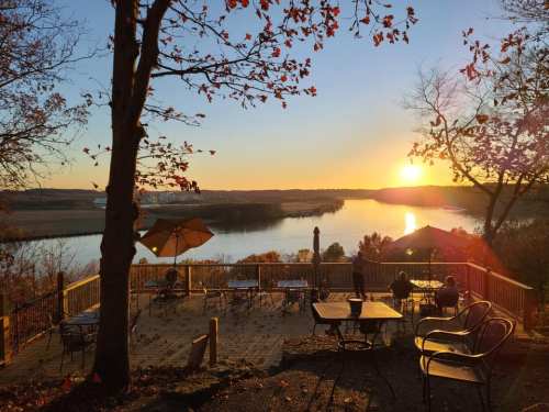 Sunset over a river viewed from a deck with tables and chairs, surrounded by autumn leaves and trees.