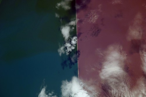 Aerial view of contrasting water colors, with a clear division between dark blue and reddish-brown sections, and clouds above.