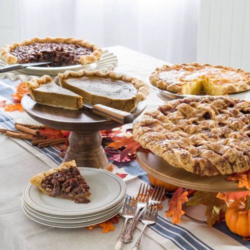 A table set with various pies, including pumpkin and pecan, surrounded by autumn leaves and utensils.