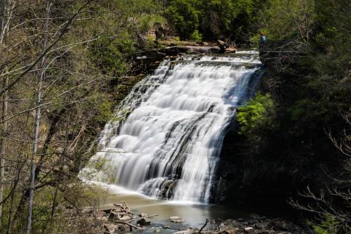 A cascading waterfall surrounded by lush greenery and rocky terrain, with a person standing near the top.
