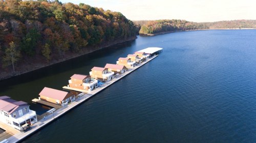 Aerial view of a dock with houseboats along a calm lake, surrounded by colorful autumn trees.