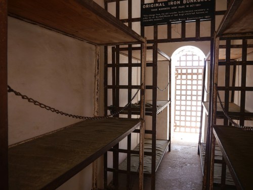 Interior of a historic prison cell with empty metal bunks and a barred door, evoking a sense of confinement.