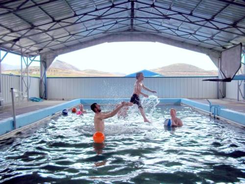 A boy jumps into a pool while another person lifts him, with others swimming and enjoying the water in the background.