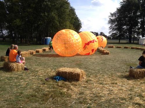Colorful inflatable balls on a grassy field, with people sitting on hay bales nearby. Trees in the background.