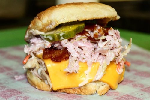 A close-up of a sandwich with pulled pork, coleslaw, cheddar cheese, pickles, and barbecue sauce on a soft bun.