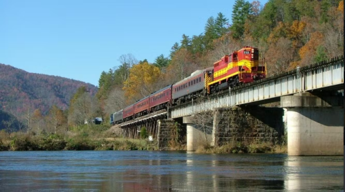 A colorful train crosses a bridge over a river, surrounded by autumn trees and mountains in the background.