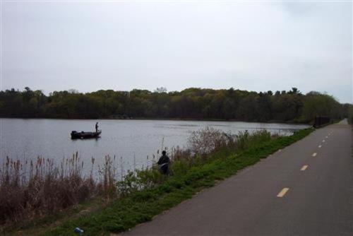 A calm lake with a fishing boat and a person fishing from the shore, surrounded by greenery and a paved path.