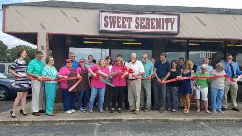 A group of people in colorful attire gather outside a store named "Sweet Serenity" for a ribbon-cutting ceremony.