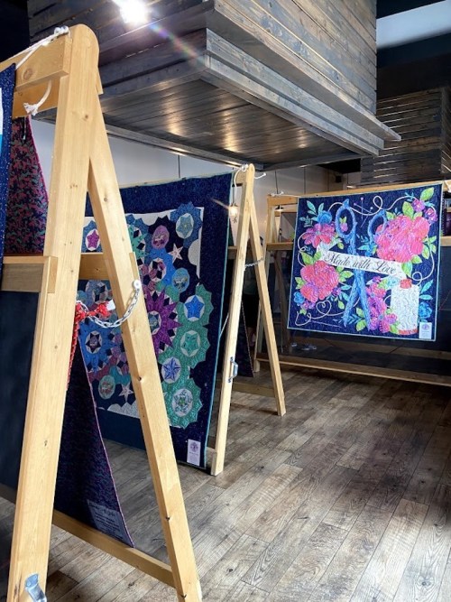 Colorful quilts displayed on wooden frames in a cozy, well-lit space with wooden flooring.