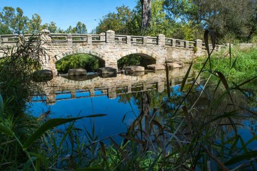 A stone bridge arches over a calm river, reflecting greenery and trees in the water below.