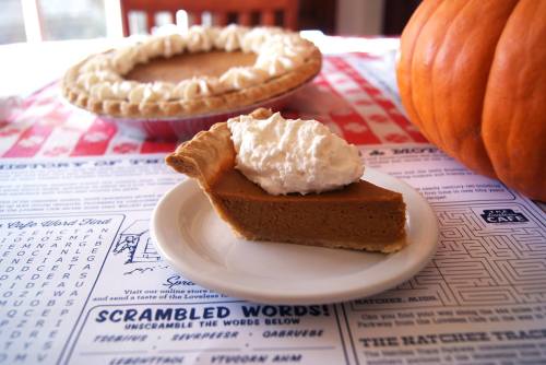 A slice of pumpkin pie topped with whipped cream on a plate, with a whole pie and a pumpkin in the background.