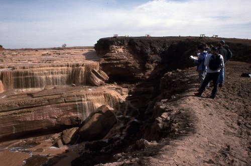 A group of people observing a series of cascading waterfalls and rocky cliffs under a clear sky.