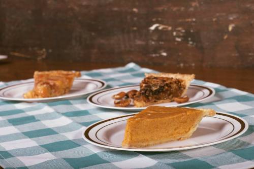 Three slices of pie on white plates: pumpkin, pecan, and a fruit pie, arranged on a checkered tablecloth.