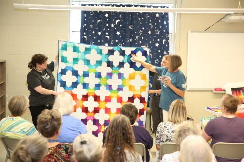 Two women hold up a colorful quilt in front of an audience seated in a classroom setting.