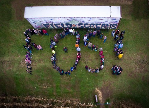 Aerial view of a large group of people forming the number "703" in a field, with a colorful mural in the background.