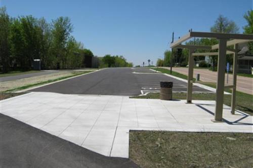 A wide, empty road lined with trees, featuring a bus stop shelter and a trash can nearby. Clear blue sky above.