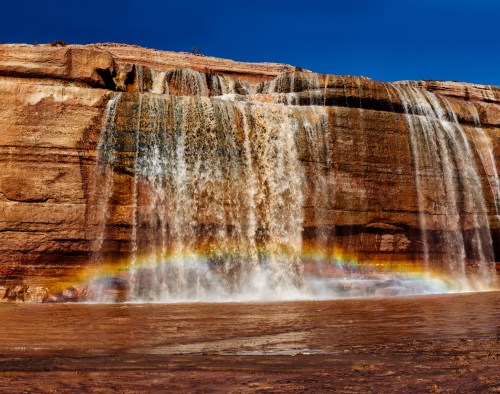 A vibrant waterfall cascades over rocky cliffs, creating a rainbow in the mist against a clear blue sky.