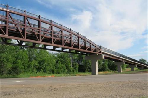 A curved pedestrian bridge with a metal frame spans over a road, surrounded by greenery and blue skies.