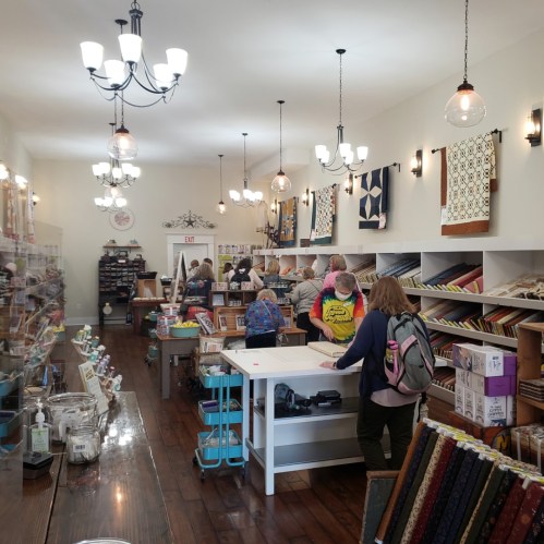 A cozy fabric store with customers browsing colorful fabrics and sewing supplies under warm lighting.
