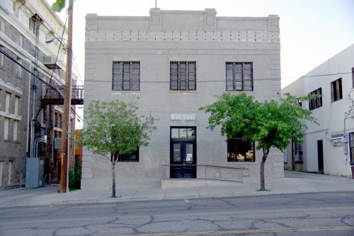 A gray, historic building with barred windows, flanked by small trees, located on a city street.