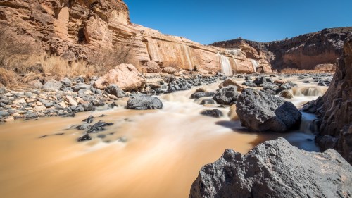 A serene river flows past rocky banks, with a waterfall cascading in the background under a clear blue sky.