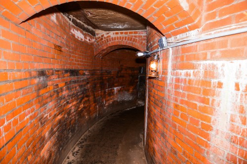 A dimly lit, curved brick tunnel with a wet floor and a light fixture on the wall.
