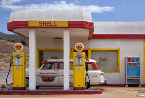 A vintage Shell gas station with classic pumps and a retro car parked outside against a scenic backdrop.