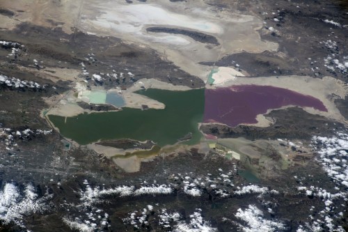 Aerial view of a colorful landscape featuring lakes in various shades of green, purple, and blue, surrounded by dry terrain.