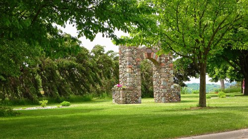A stone archway surrounded by lush green trees and grass, with colorful flowers at its base.