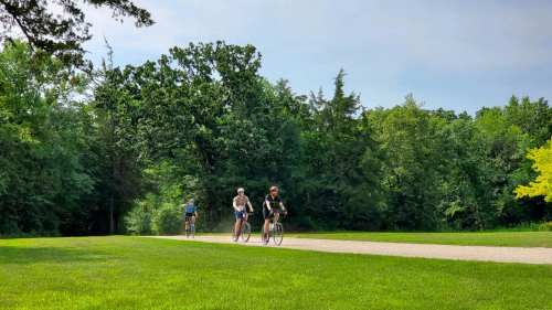 Three cyclists ride along a gravel path surrounded by lush green trees and grass under a clear blue sky.