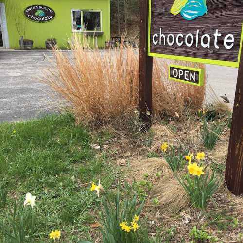 A sign for a chocolate shop reads "chocolate" with "OPEN" below, surrounded by greenery and blooming flowers.