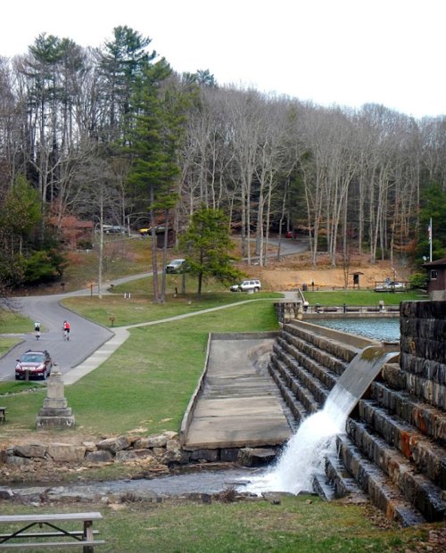 A serene landscape featuring a waterfall cascading over stone steps, surrounded by trees and a pathway with parked cars.