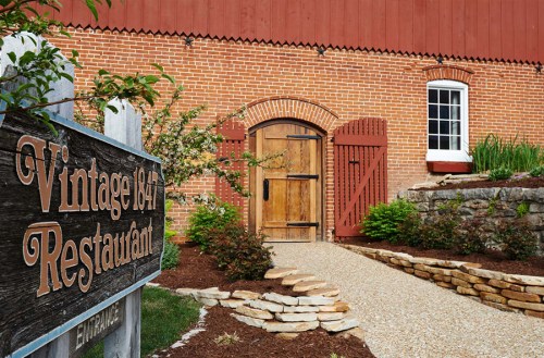 A rustic restaurant entrance with a wooden door, surrounded by greenery and stone pathways, featuring a vintage sign.