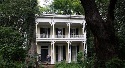 A two-story white house with a porch, surrounded by trees and greenery. Two people are visible near the entrance.
