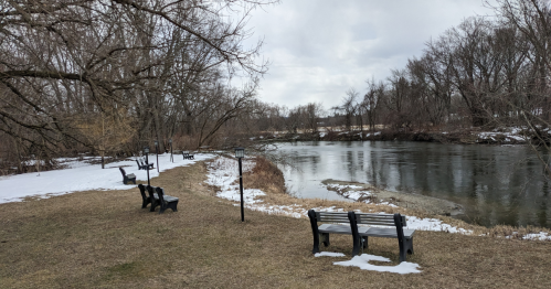 A serene riverside scene with benches along a snowy bank and bare trees under a cloudy sky.