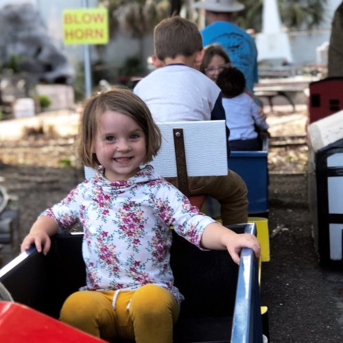 A young girl in a colorful outfit sits in a toy train, smiling, with children playing in the background.
