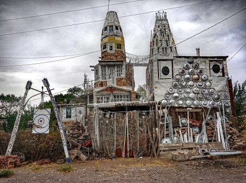 An eclectic, abandoned building with towers, adorned with various metal objects and sculptures, set against a cloudy sky.