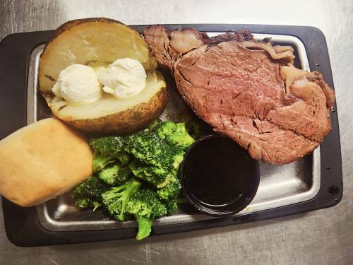 A plate with prime rib, a baked potato with butter, steamed broccoli, a roll, and a side of sauce.