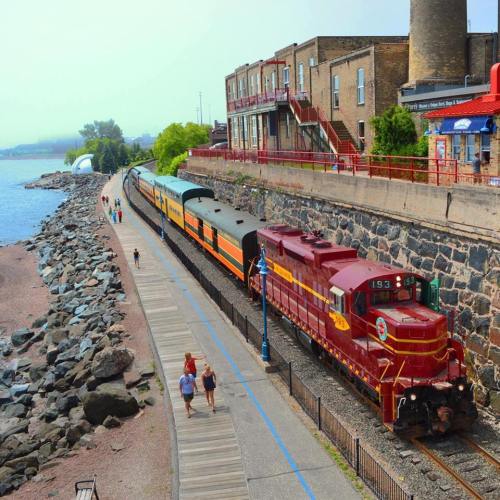 A vintage train parked along a scenic waterfront, with people walking on a path beside the water and buildings in the background.