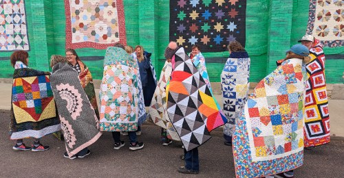 A group of people wrapped in colorful quilts stands in front of a mural featuring various quilt patterns.