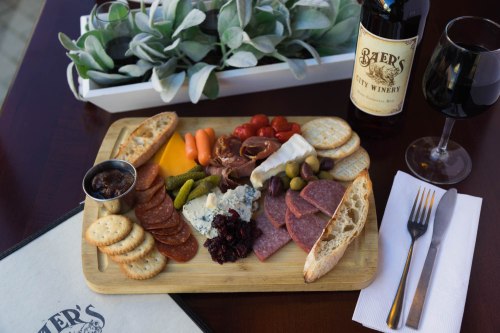 A wooden platter with assorted meats, cheeses, crackers, olives, and vegetables, alongside a bottle of wine and a glass.