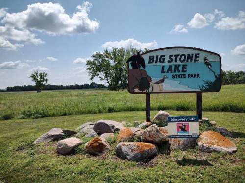 Sign for Big Stone Lake State Park with a grassy field and trees in the background, featuring colorful rocks in the foreground.