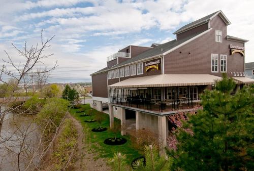 A brown building with a patio overlooks a river, surrounded by greenery and under a partly cloudy sky.