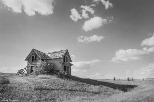 A weathered, abandoned house on a grassy hill under a cloudy sky, captured in black and white.