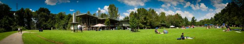 A panoramic view of a park with people relaxing on the grass, trees, and a building in the background under a blue sky.